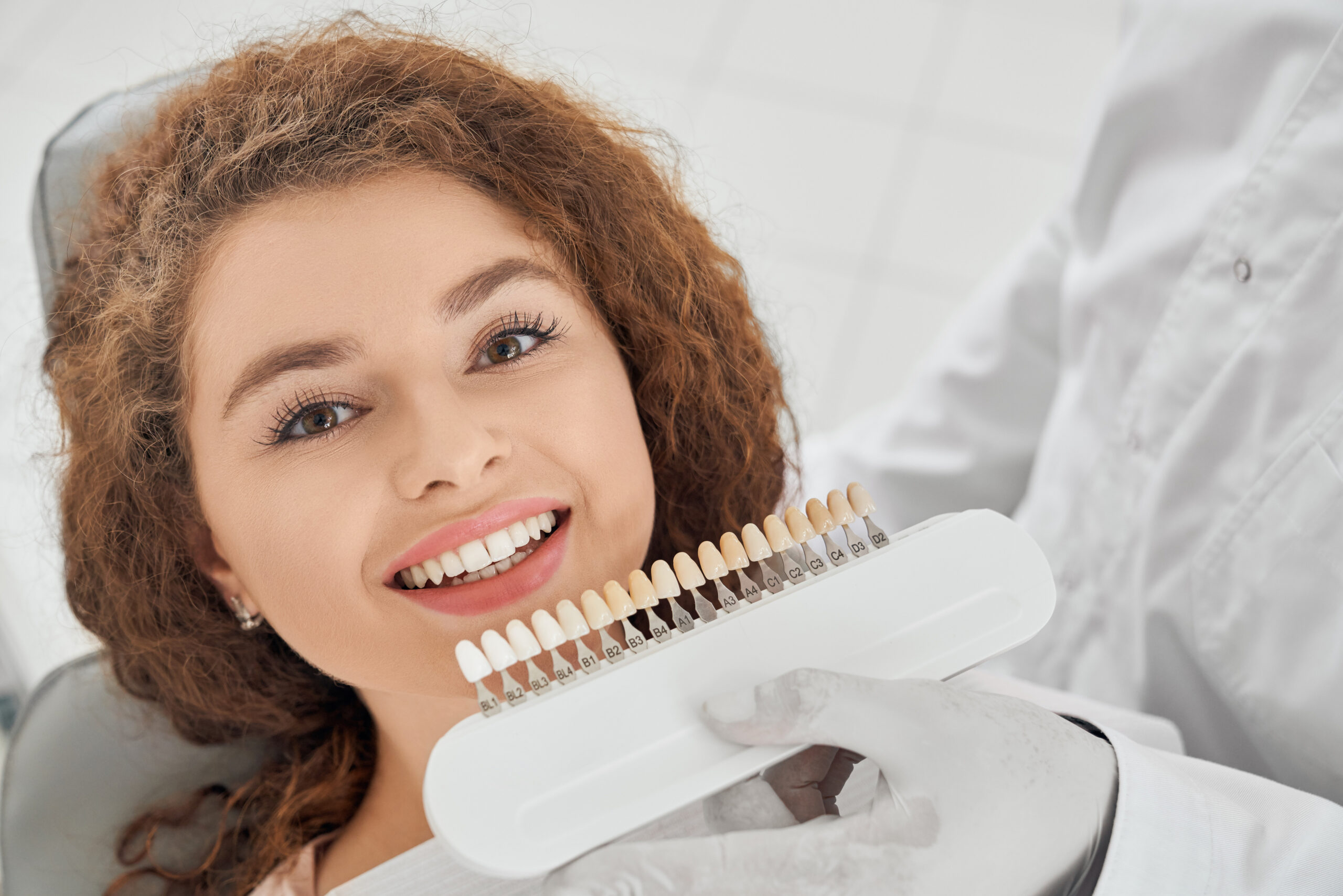 Closeup of a female on a dental char, while male dentist keeping in hands teeth color range. Girl doing whitening procedure in dental office.